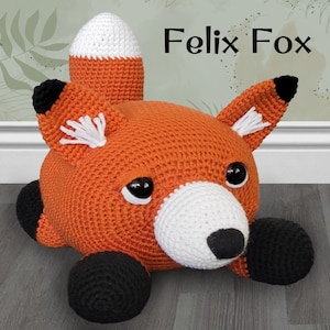 May include: A crocheted orange fox plush toy with black eyes, a white nose, and black paws. The fox is lying on its belly with its tail curled up behind it. The text "Felix Fox" is written in black letters above the fox.