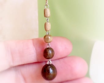 May include: A long wooden bead earring with a silver hook. The earring has a mix of brown and light brown beads.