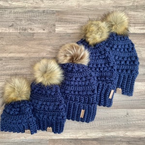 May include: Four navy blue crocheted beanies with faux fur pom poms in various sizes. The beanies are lined up from largest to smallest.