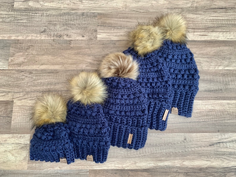 May include: Four navy blue crocheted beanies with faux fur pom poms in various sizes. The beanies are lined up from largest to smallest.