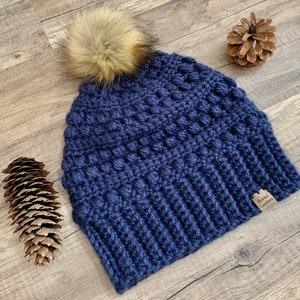 May include: A navy blue crocheted beanie with a faux fur pom pom on top. The beanie has a textured stitch pattern and a brown leather tag that says "Riley Crafts".