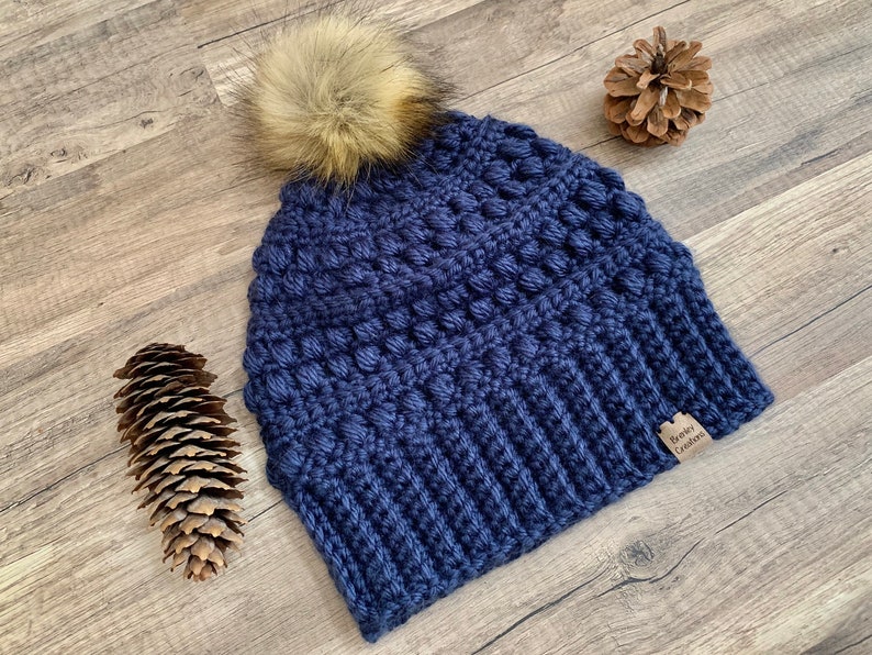 May include: A navy blue crocheted beanie with a faux fur pom pom on top. The beanie has a textured stitch pattern and a brown leather tag that says "Riley Crafts".