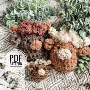 May include: Three crocheted stuffed animal Highland cows in brown, tan, and cream colors. The cows are sitting on a brown and white patterned fabric. The text "PDF PATTERN" is visible in the image.