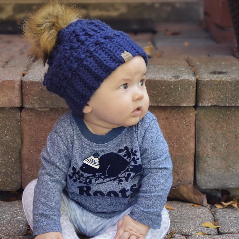 May include: A baby wearing a blue knitted hat with a brown pom pom and a blue long-sleeved shirt with a beaver graphic and the word "Roots" on it.