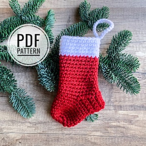 May include: A red and white crocheted Christmas stocking with a white cuff and a loop for hanging. The stocking is on a wooden background with green pine branches.