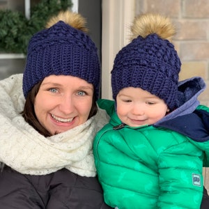 May include: A woman and a child wearing matching navy blue knitted hats with faux fur pom-poms. The woman is wearing a white scarf and a black jacket. The child is wearing a green puffer jacket with a dinosaur print.