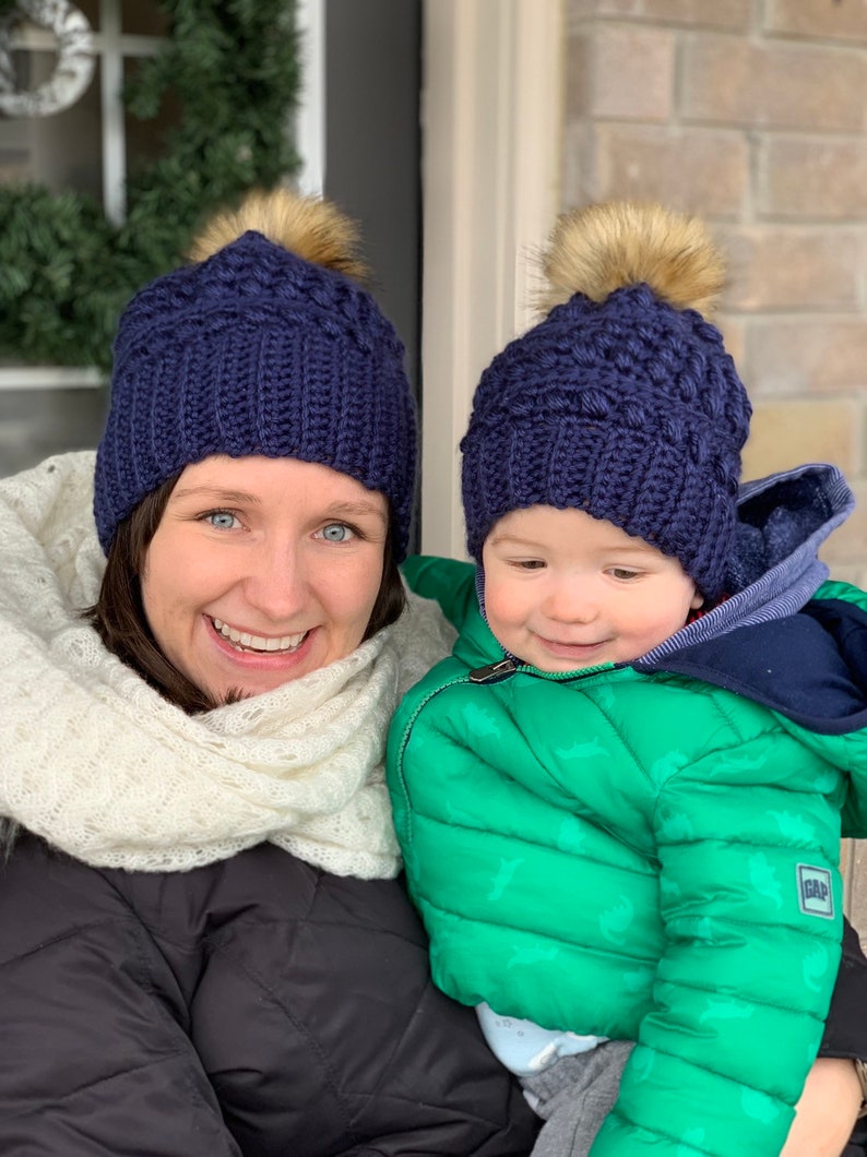 May include: A woman and a child wearing matching navy blue knitted hats with faux fur pom-poms. The woman is wearing a white scarf and a black jacket. The child is wearing a green puffer jacket with a dinosaur print.