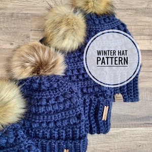 May include: Four navy blue crochet winter hats with faux fur pom poms. The hats have a textured stitch pattern and a brown leather tag that says "Bradley Creations". The image includes the text "Winter Hat Pattern".