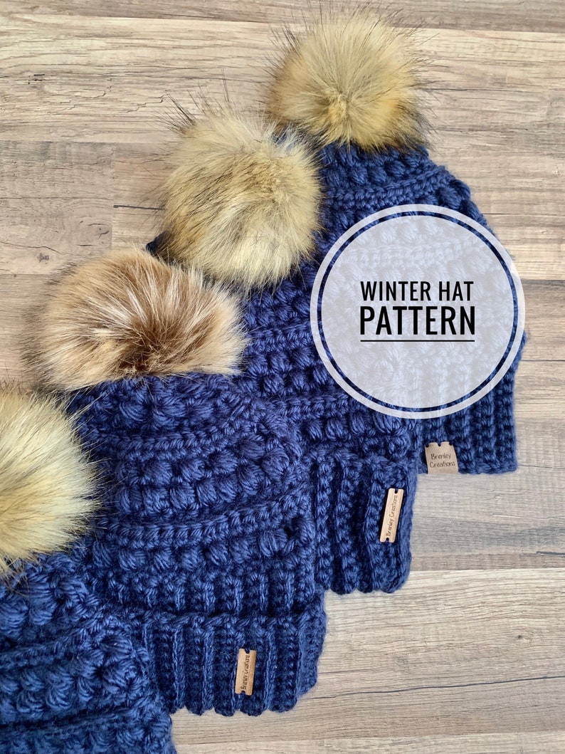 May include: Four navy blue crochet winter hats with faux fur pom poms. The hats have a textured stitch pattern and a brown leather tag that says "Bradley Creations". The image includes the text "Winter Hat Pattern".