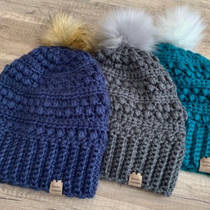 May include: Three crocheted beanies with pom poms in navy blue, grey, and teal. Each beanie has a small brown tag that reads "Snuggly Creations".