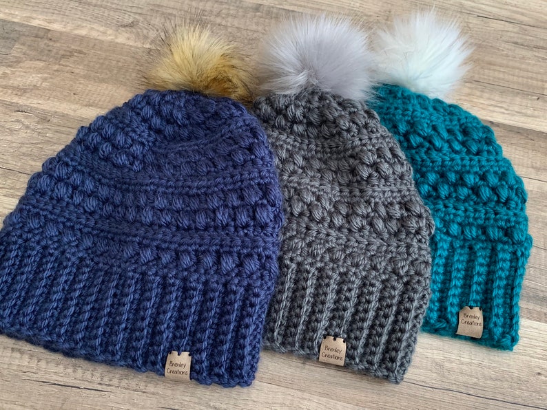 May include: Three crocheted beanies with pom poms in navy blue, grey, and teal. Each beanie has a small brown tag that reads "Snuggly Creations".