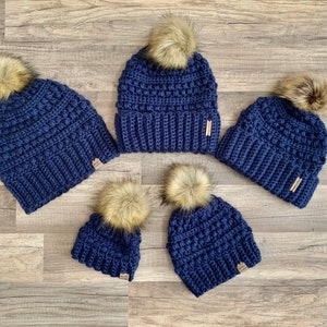 May include: Set of four navy blue crocheted beanies with faux fur pom poms. The beanies are in different sizes.
