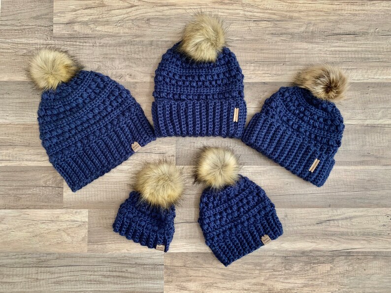 May include: Set of four navy blue crocheted beanies with faux fur pom poms. The beanies are in different sizes.