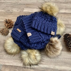 May include: A navy blue crocheted hat and scarf set with faux fur pom poms. The set has a small brown tag that says "Sewn Creations".