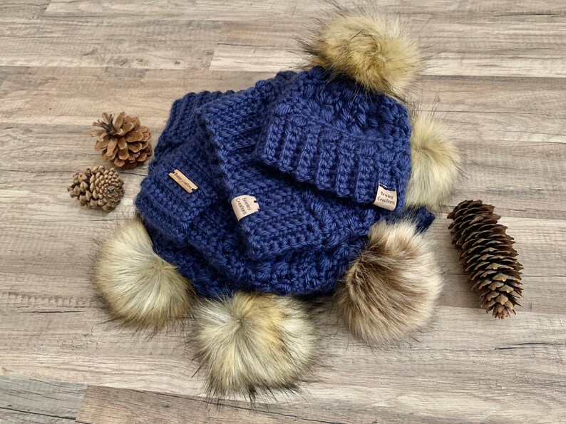 May include: A navy blue crocheted hat and scarf set with faux fur pom poms. The set has a small brown tag that says "Sewn Creations".