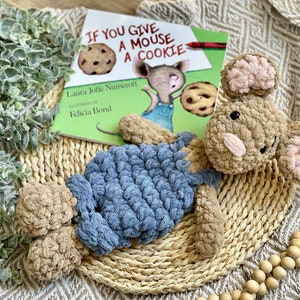 May include: A brown and blue crocheted mouse plush toy with pink ears. The toy is lying on a woven basket with a copy of the children's book "If You Give a Mouse a Cookie" in the background.