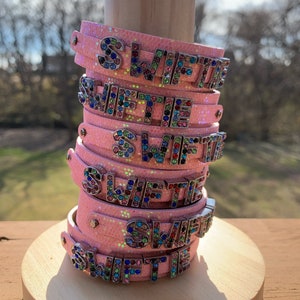 May include: Stack of pink glitter bracelets with the name "SWIFTIE" spelled out in silver letters encrusted with colorful rhinestones. The bracelets are stacked on a wooden display stand.
