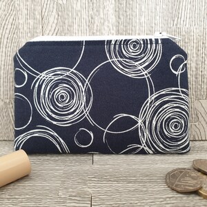 May include: Dark blue coin purse with a white spiral design. The small pouch has a white zip. The fabric design features white swirling circles on a navy blue background. A practical accessory.
