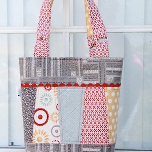 Op de afbeelding: Een grijze en witte patchwork-tas met rood-witte gepatroneerde hengsels. De tas heeft een rood-witte bies en een label met de tekst "Lily Patch Quilts".