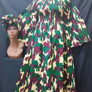 May include: A long, flowing dress and matching head wrap in a camouflage pattern of green, burgundy, and tan. The dress features a gathered bodice and wide sleeves. The head wrap is styled in a turban shape. The outfit is displayed on a mannequin.