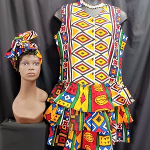 May include: A sleeveless dress with a colorful geometric pattern in shades of red, yellow, blue, green, and black. The dress has a ruffled skirt and a button-up front. A matching head wrap is also included.