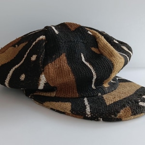 May include: A patterned cap in black, brown, and white. The cap features a visor and a rounded crown. The fabric has a woven texture, with a design of irregular shapes and stripes. The color palette is neutral, making it a versatile accessory.