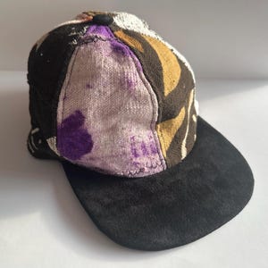 May include: A baseball cap with a black suede bill. The crown features a patchwork design with shades of brown, purple, and tan. The cap has a classic six-panel construction and a rounded top.