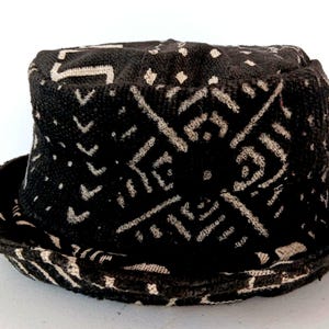 May include: A black and white patterned hat with a rounded crown and a brim. The hat features a repeating geometric design in white against a black background. The brim is also patterned with the same design.
