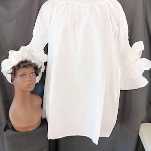 May include: A white blouse with a high, ruffled neckline and long sleeves with ruffled cuffs. The blouse is displayed on a mannequin. A white head wrap is on a mannequin head to the left of the blouse. The background is black.
