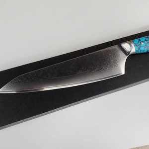 May include: A black chef's knife with a turquoise and black patterned handle. The knife is on a black display box.