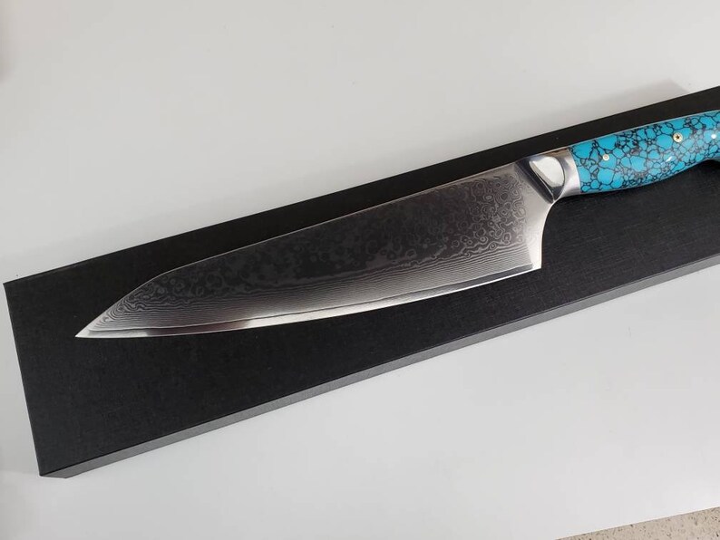 May include: A black chef's knife with a turquoise and black patterned handle. The knife is on a black display box.