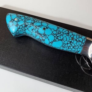 May include: A knife handle with a turquoise and black patterned design. The handle is made of a material that resembles turquoise.