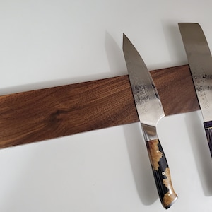 May include: A wooden magnetic knife holder with two knives attached. The holder is made of dark brown wood and has a smooth finish. The knives have different coloured handles, one is black and the other is purple.
