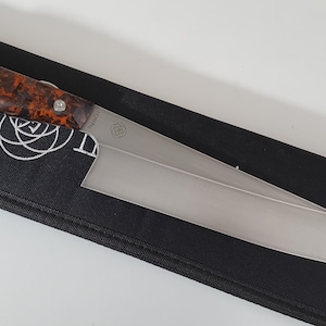 Könnte beinhalten: Ein Edelstahl-Kochmesser mit einem braun-orange gesprenkelten Griff, das auf einer schwarzen Scheide liegt. Das Messer hat eine breite Klinge und ein Logo in der Nähe des Griffs.