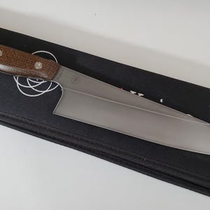 Puede incluir: Un cuchillo de chef de acero inoxidable con mango de madera marrón, sobre un estuche negro con cremallera. El cuchillo tiene una hoja ancha y rectangular y está diseñado para uso profesional. El mango está asegurado con tres remaches.