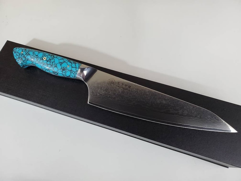 May include: A blue and silver chef's knife with a turquoise patterned handle. The knife is resting on a black surface.