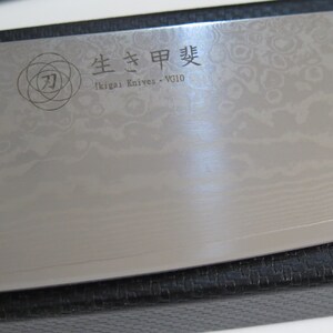 May include: A close-up of a silver chef's knife with a black handle. The knife has a decorative pattern on the blade and the text "生きた甲斐" (Ikigai Knives - VG10) is engraved on the blade.