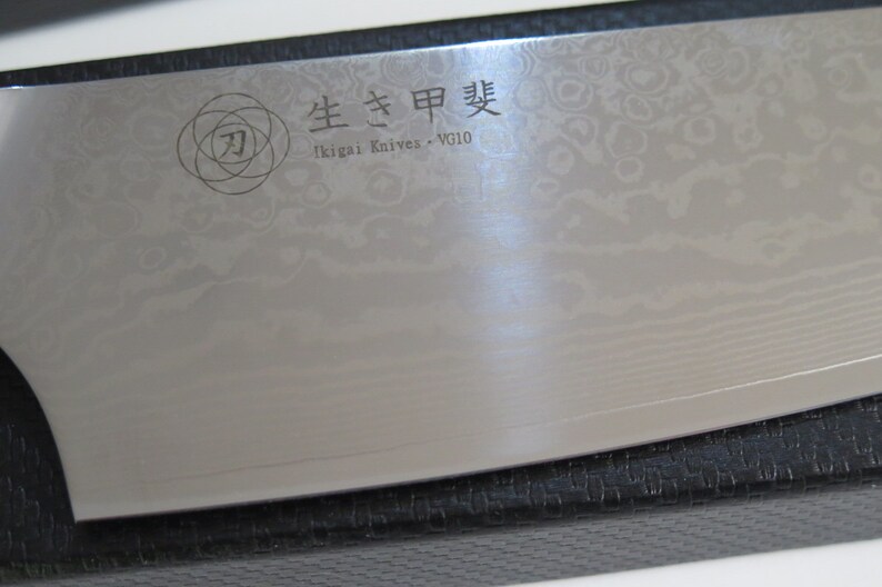 May include: A close-up of a silver chef's knife with a black handle. The knife has a decorative pattern on the blade and the text "生きた甲斐" (Ikigai Knives - VG10) is engraved on the blade.