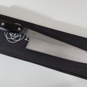 Puede incluir: Un cuchillo de chef de acero inoxidable con mango negro y tres remaches, sobre una funda negra con cremallera. El cuchillo tiene una hoja ancha y una punta afilada. La funda tiene un logotipo blanco.