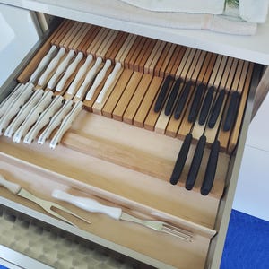May include: A kitchen drawer organizer made of light wood, featuring multiple slots holding a collection of cutlery. The set includes knives with white handles, knives with black handles, and two metal carving forks with white handles.