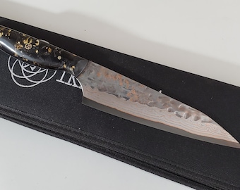 May include: A chef's knife with a dark handle inlaid with gold flecks rests on a black sheath. The blade has a textured, patterned surface. The knife is designed for kitchen use.
