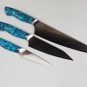 May include: Set of three kitchen knives with turquoise patterned handles. The largest knife has a black blade with a textured finish. The smaller knives have silver blades.
