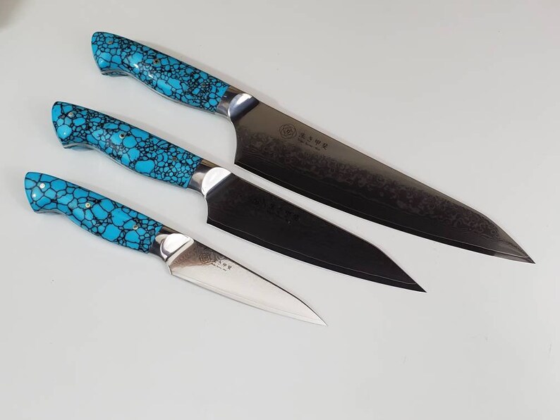 May include: Set of three kitchen knives with turquoise patterned handles. The largest knife has a black blade with a textured finish. The smaller knives have silver blades.