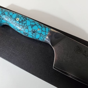 May include: A turquoise and black patterned handle with gold accents on a Japanese chef's knife with a Damascus steel blade. The blade has a rose pattern and the text "生粋甲斐" (pronounced "Seisui Kai") and "Japan Damascus VG10" is engraved on the blade.
