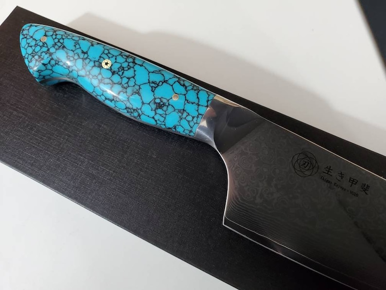 May include: A turquoise and black patterned handle with gold accents on a Japanese chef's knife with a Damascus steel blade. The blade has a rose pattern and the text "生粋甲斐" (pronounced "Seisui Kai") and "Japan Damascus VG10" is engraved on the blade.