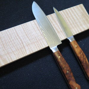 Peut inclure: Deux couteaux de cuisine avec des manches en bois sont fixés à une bande magnétique en bois clair avec un motif de grain ondulé.