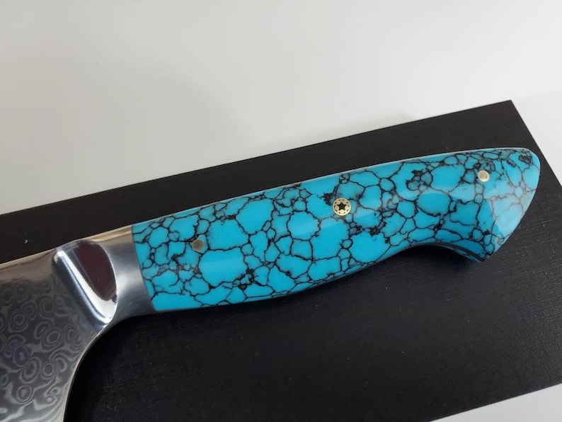 May include: A turquoise and black patterned knife handle with gold accents. The handle is made of a material that resembles turquoise stone.