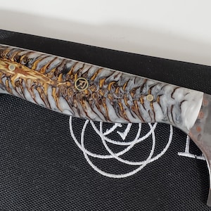 May include: Close-up of a chef's knife with a decorative handle. The handle features a white, brown, and gold pattern. The blade has a textured surface with a floral design and Japanese characters.