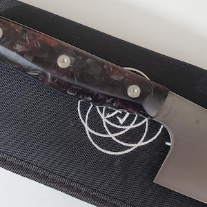 Könnte beinhalten: Ein Edelstahl-Kochmesser mit einem dunklen, gemusterten Griff, der mit drei silbernen Nieten befestigt ist. Die Klinge ist teilweise sichtbar und liegt auf einer schwarzen Stoffoberfläche mit weißem Design. Küchenmesser.