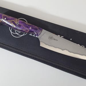 Puede incluir: Un cuchillo con un patrón morado y dorado con una hoja de acero inoxidable y una funda de cuero negro. La hoja tiene un borde ondulado y el mango tiene un diseño de rosa con el texto "20CV".